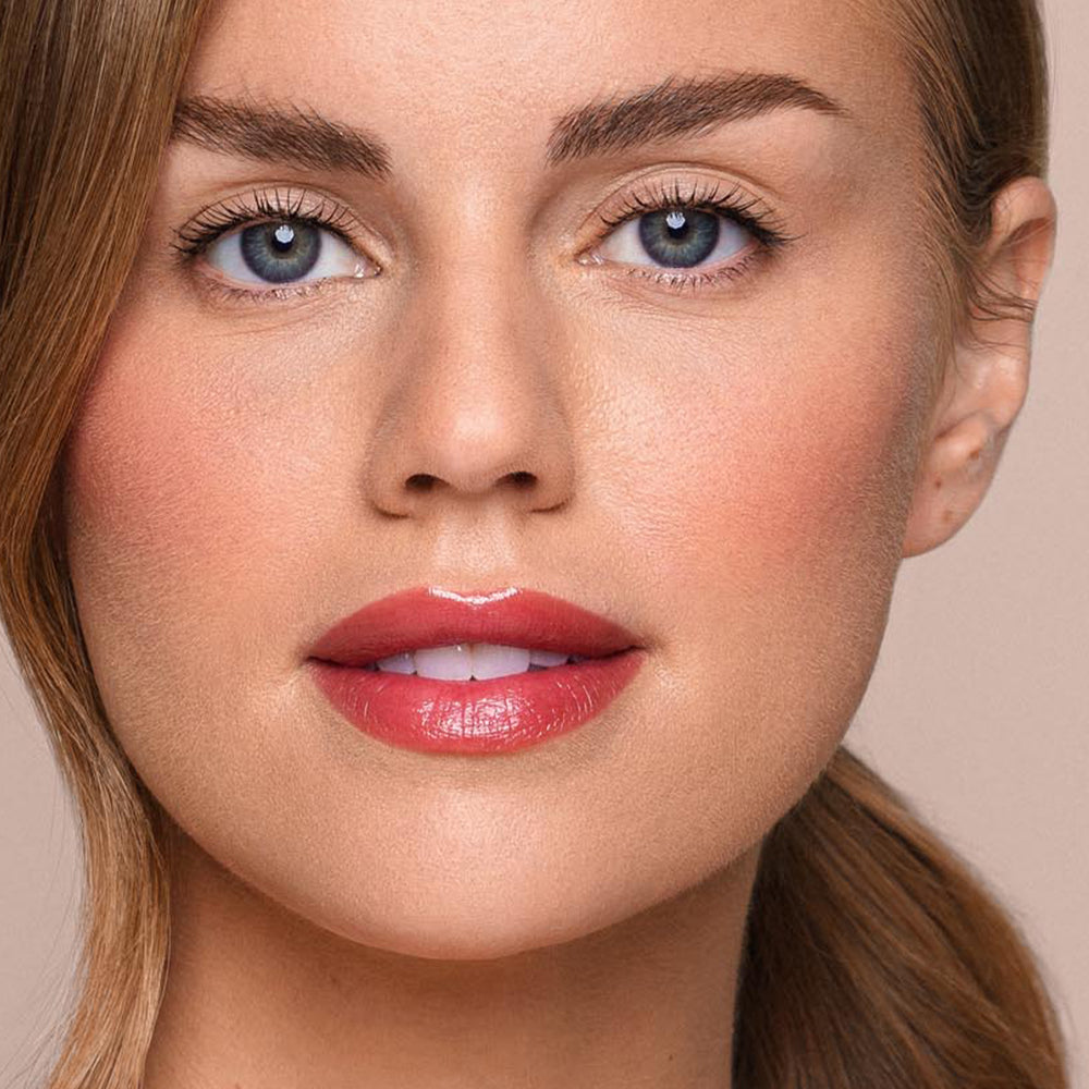 A close-up of an individual with fair skin, light blue eyes, and full, glossy pink lips enhanced by Jouer Cosmetics' Essential Shine Balm. They boast well-groomed eyebrows and light makeup applied using paraben-free products. Their brown hair is pulled back, revealing their face against a plain background.
