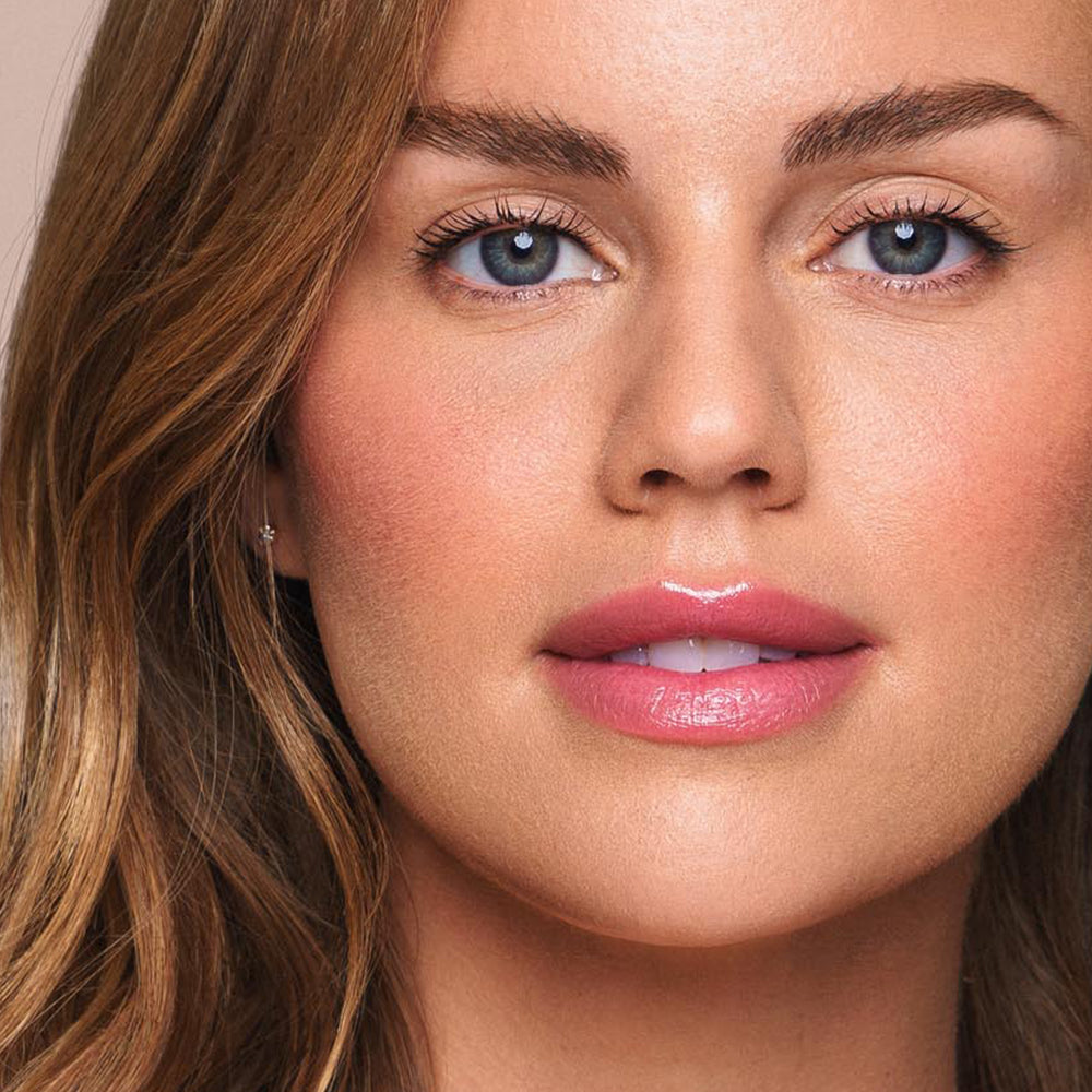 A close-up of a woman with wavy light brown hair and blue eyes showcases her natural, radiant makeup featuring defined eyebrows, long lashes, rosy cheeks, and glossy pink lips. Her serene expression beautifully highlights the cruelty-free and vegan Essential Shine Balm by Jouer Cosmetics used for her look.