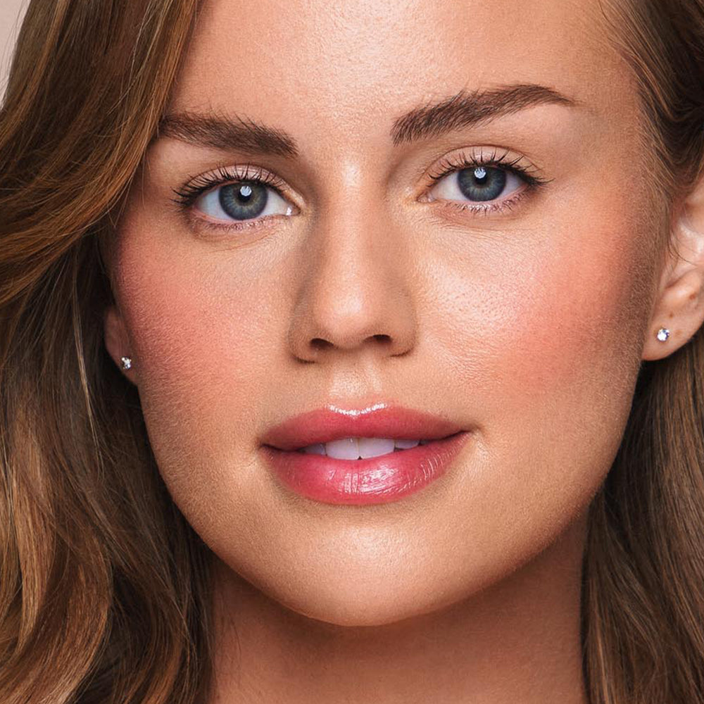 A close-up of a woman with light brown hair and blue eyes. Her cheeks are subtly rosy, and she wears light, paraben-free makeup with glossy pink lips using Jouer Cosmetics' Essential Shine Balm. Small diamond stud earrings adorn her ears. She looks directly at the camera with a neutral expression.