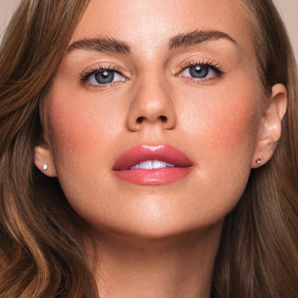 A close-up of a woman with light skin, long brown hair, blue eyes, and well-defined eyebrows. She has a subtle rosy blush, glossy pink lips thanks to the Essential Shine Balm by Jouer Cosmetics, and is wearing small stud earrings. All products used are cruelty-free and paraben-free. The background is neutral.