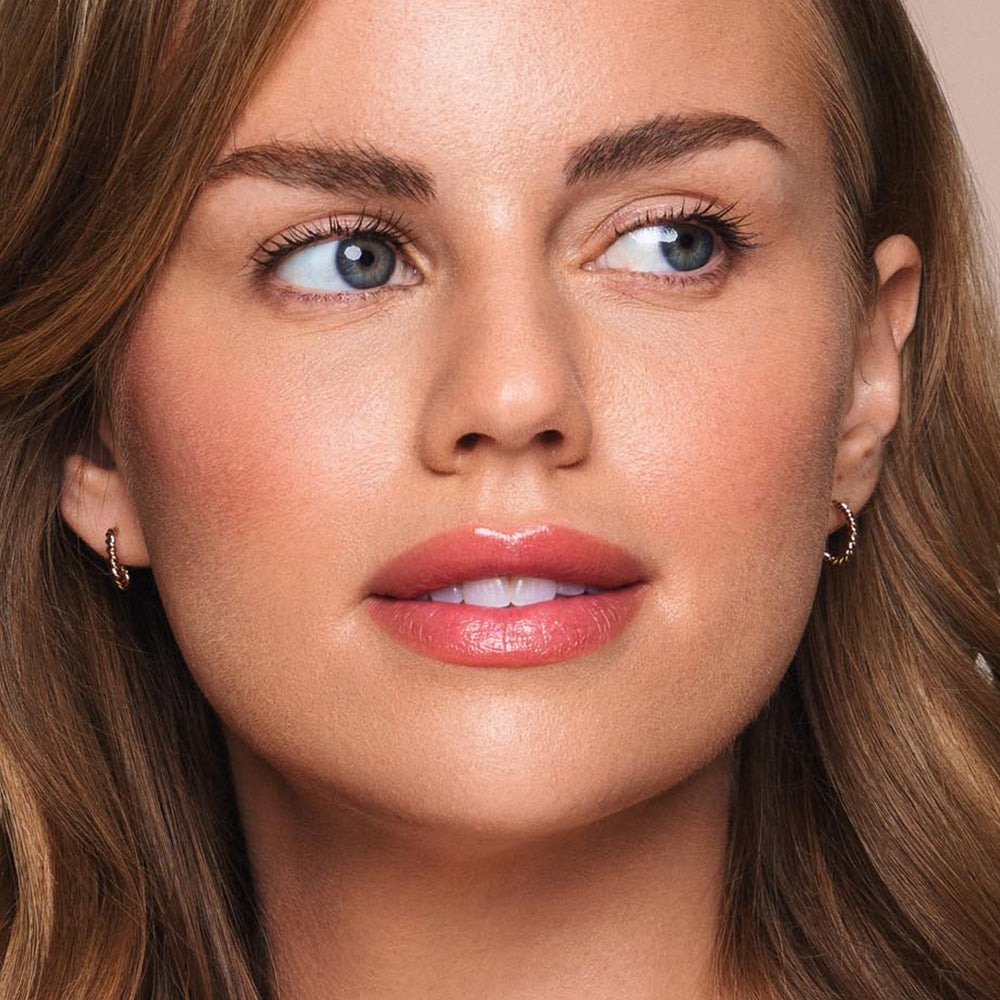 A woman with wavy brown hair and blue eyes looks slightly to her right. She has a natural makeup look featuring pink lipstick, subtle blush, and well-defined eyebrows, all crafted from vegan products by Jouer Cosmetics' Essential Shine Balm. She's wearing small hoop earrings and exudes a calm expression against a plain background.