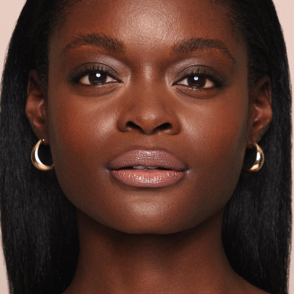 Close-up of a woman with smooth, dark skin and straight, long black hair. She is wearing gold hoop earrings. Her facial expression is calm, and she is looking directly at the camera. This moment captures the essence of a cruelty-free and vegan lifestyle, embodying the natural glow provided by Jouer Cosmetics' Essential Shine Balm.