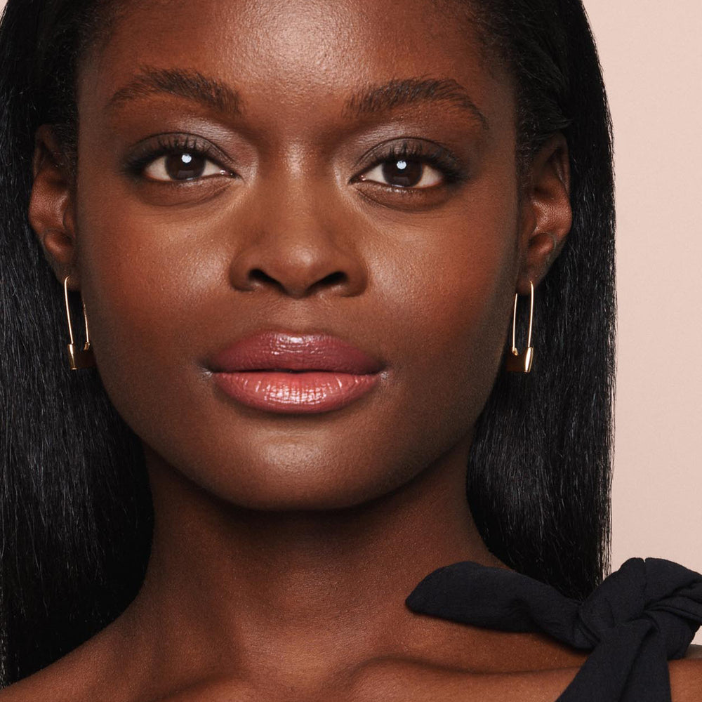 Close-up of a person with smooth, dark skin, wearing gold rectangular hoop earrings, glossy pink Essential Shine Balm lipstick from Jouer Cosmetics that is vegan and paraben-free, and black attire with a bow on one shoulder. They have long, straight, dark hair and are looking directly at the camera with a neutral expression.