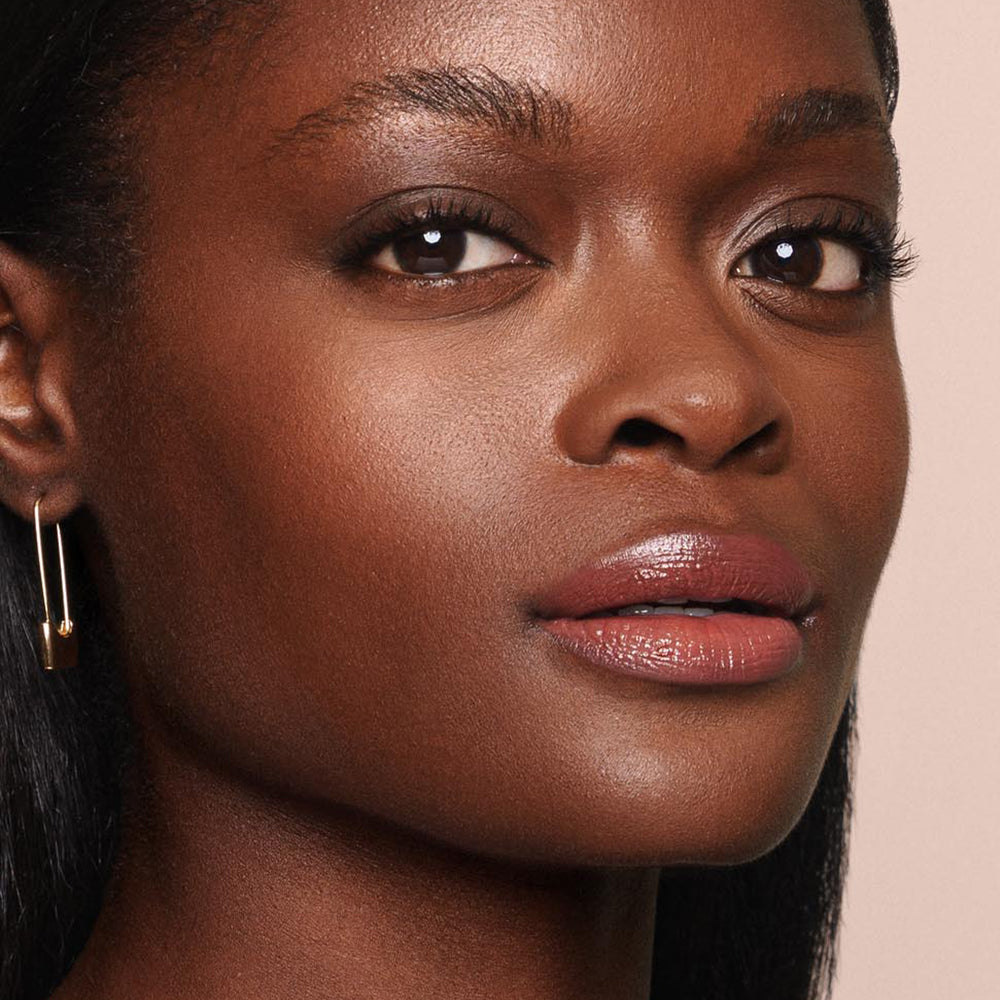 A close-up of a person with dark skin, looking slightly to the side. They have well-defined eyebrows, subtle eye makeup, and glossy lips enhanced by Jouer Cosmetics' Essential Shine Balm—both vegan and cruelty-free. They wear a single earring on their left ear, their expression serene against a soft, neutral background.