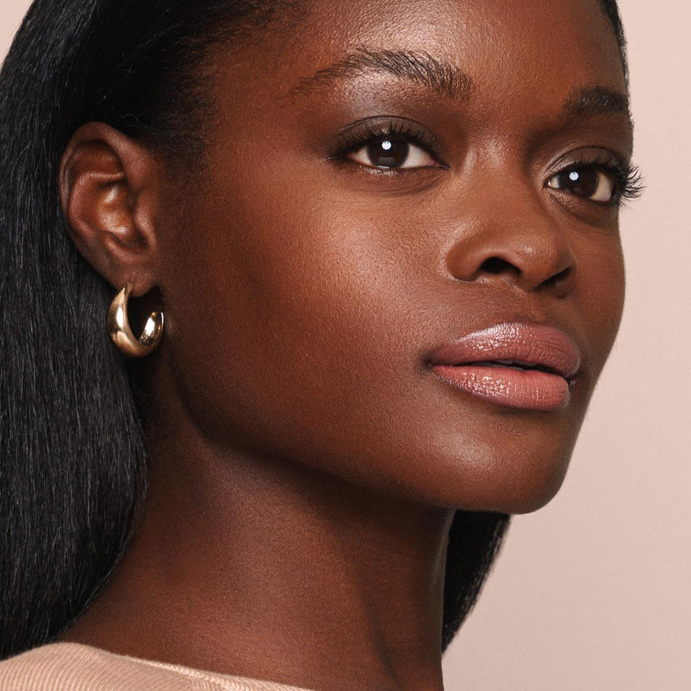 Close-up portrait of a person with smooth, dark skin wearing gold hoop earrings and neutral-colored makeup from Jouer Cosmetics' Essential Shine Balm. Their hair is straight and long, cascading over their shoulders. The background is a soft beige, creating a warm and polished look.