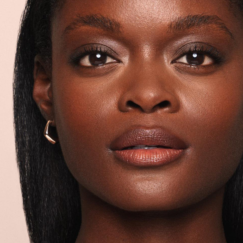 Close-up of a person with smooth, radiant brown skin, wearing a subtle, paraben-free makeup look featuring glossy lips courtesy of Jouer Cosmetics' Essential Shine Balm and slightly shimmering eyeshadow. They have straight, dark hair and are wearing a small, simple hoop earring in one ear. The background is neutral.