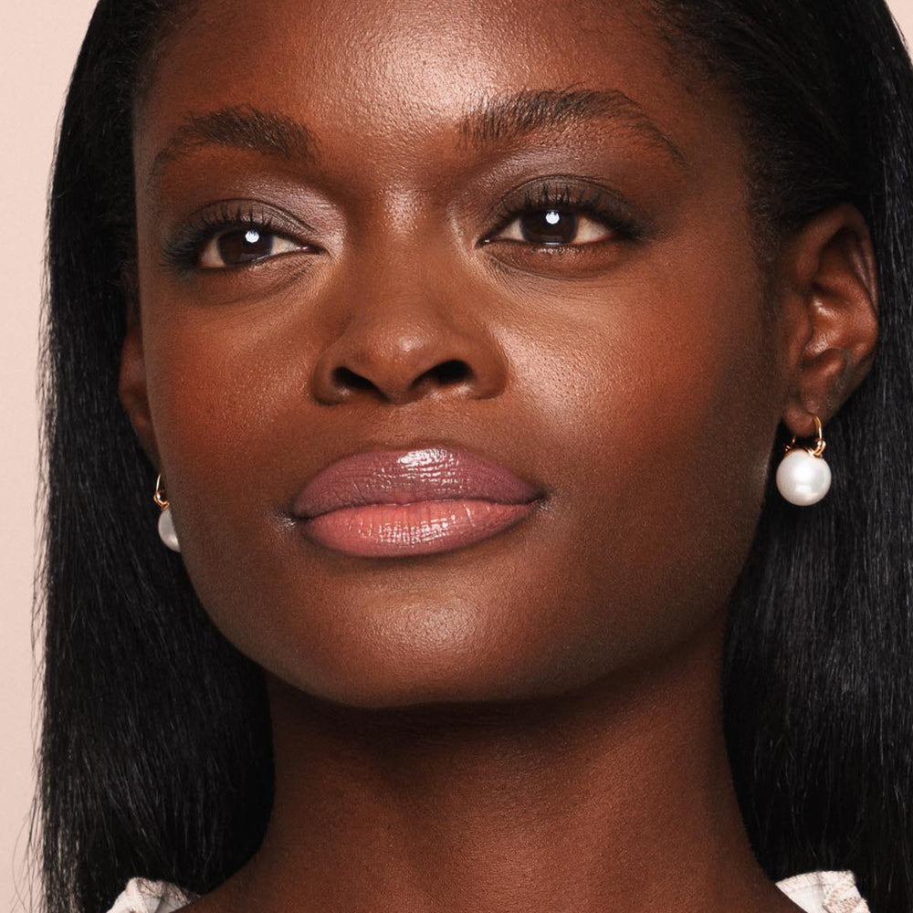 A close-up of a woman with a calm expression. She has smooth, radiant, paraben-free skin, glossy pink Essential Shine Balm from Jouer Cosmetics on her lips, subtle eye makeup, and is wearing pearl drop earrings. Her straight black hair is parted in the middle and she is dressed in a white top.