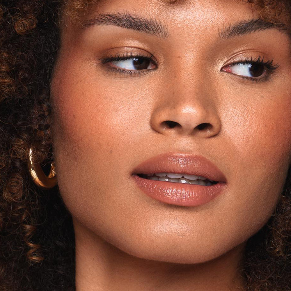 A close-up of a person's face showing smooth, glowing skin with a natural makeup look, achieved with Jouer Cosmetics' Essential Shine Balm. They have curly hair framing their face, gold hoop earrings, and a neutral expression with slightly parted lips and a soft gaze looking off to the side—all achieved using vegan, cruelty-free products.