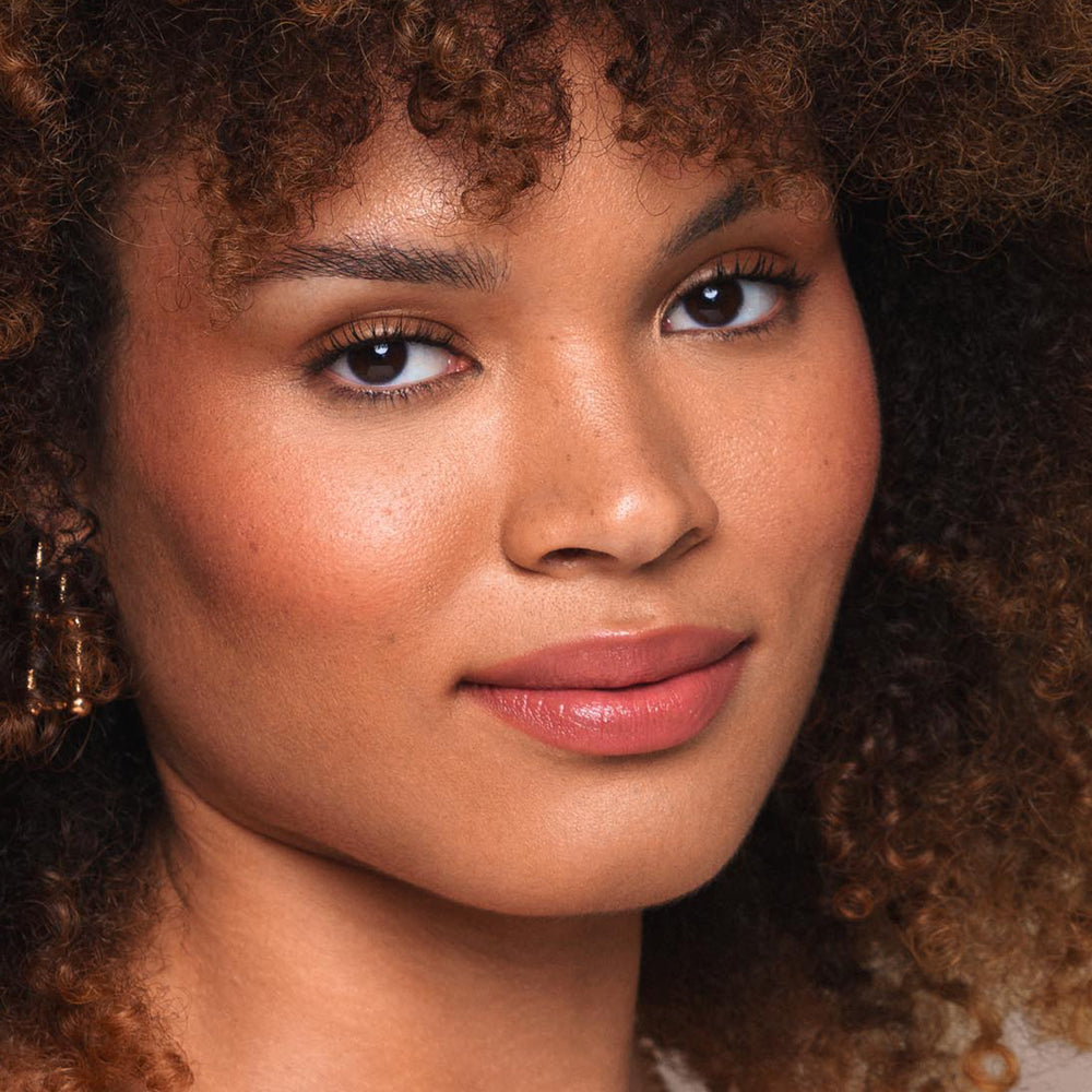 Close-up of a person with curly brown hair, smooth skin, and a subtle smile. They have natural makeup featuring Jouer Cosmetics' Essential Shine Balm, which is cruelty-free and paraben-free. Wearing earrings, they look directly at the camera in a well-lit setting.