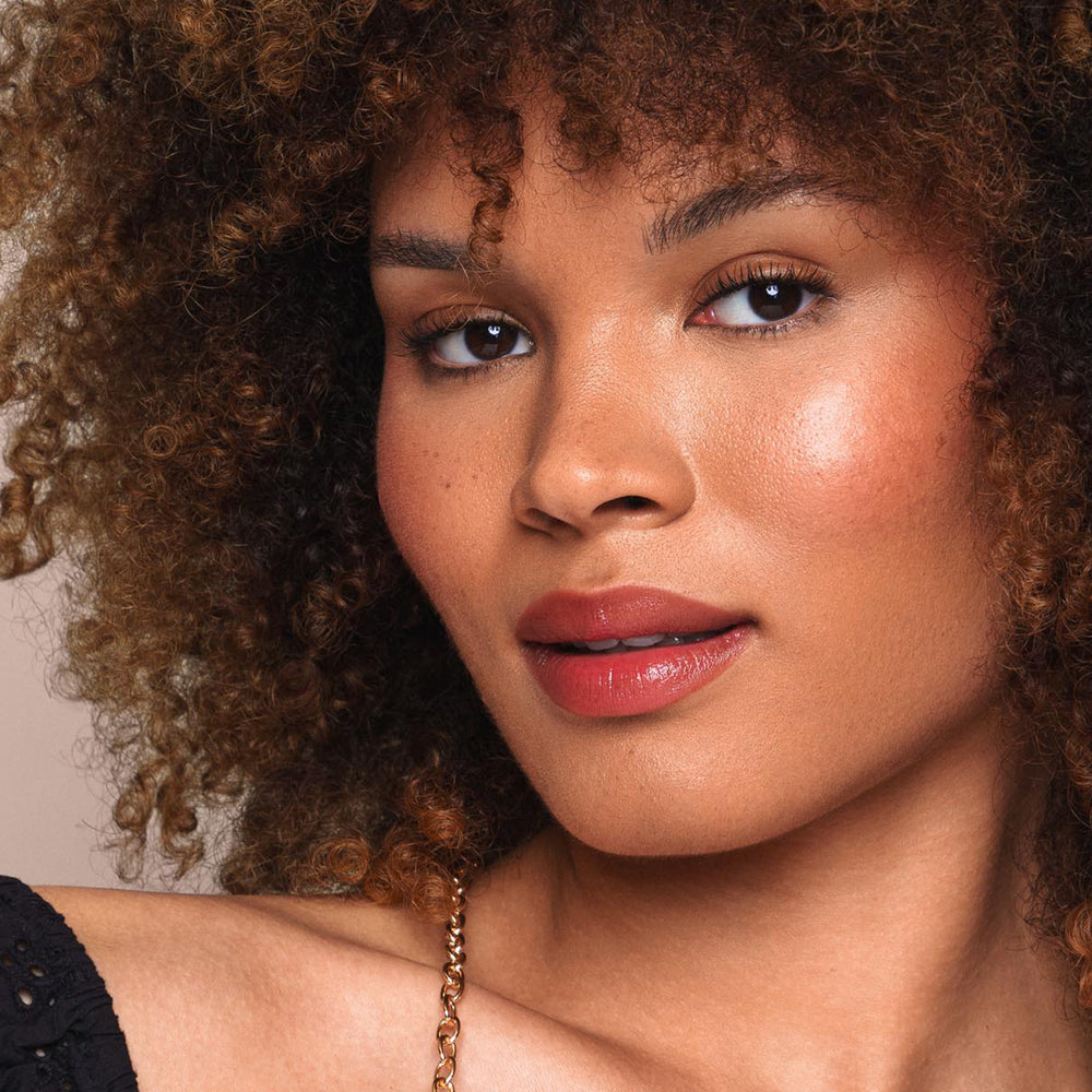 A close-up of a person with medium brown skin and voluminous, curly hair showcasing their subtle, cruelty-free makeup by Jouer Cosmetics. Their glistening lips feature the Essential Shine Balm, complementing their softly highlighted cheeks. They exude a calm and confident expression while dressed in a black top.