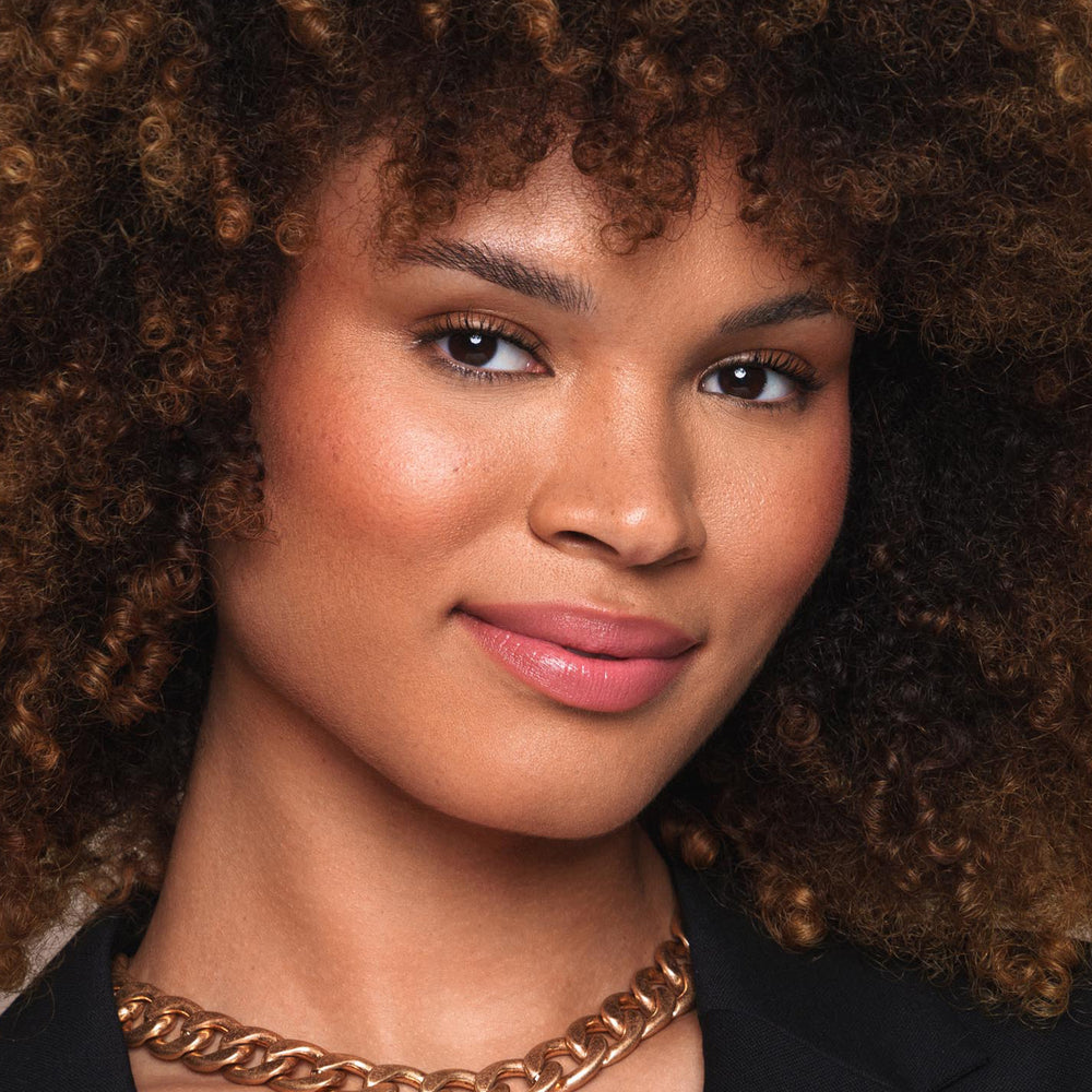 A person with a glowing complexion and curly, voluminous hair poses for a close-up portrait. They have a subtle smile, wear a gold chain necklace, and are dressed in black attire. Their natural makeup, enhanced by Jouer Cosmetics' Essential Shine Balm which is cruelty-free and vegan, subtly highlights their features.