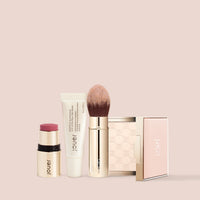 Jouer Les Must-Haves makeup set with lipstick, foundation, highlighter and brush on pink background
