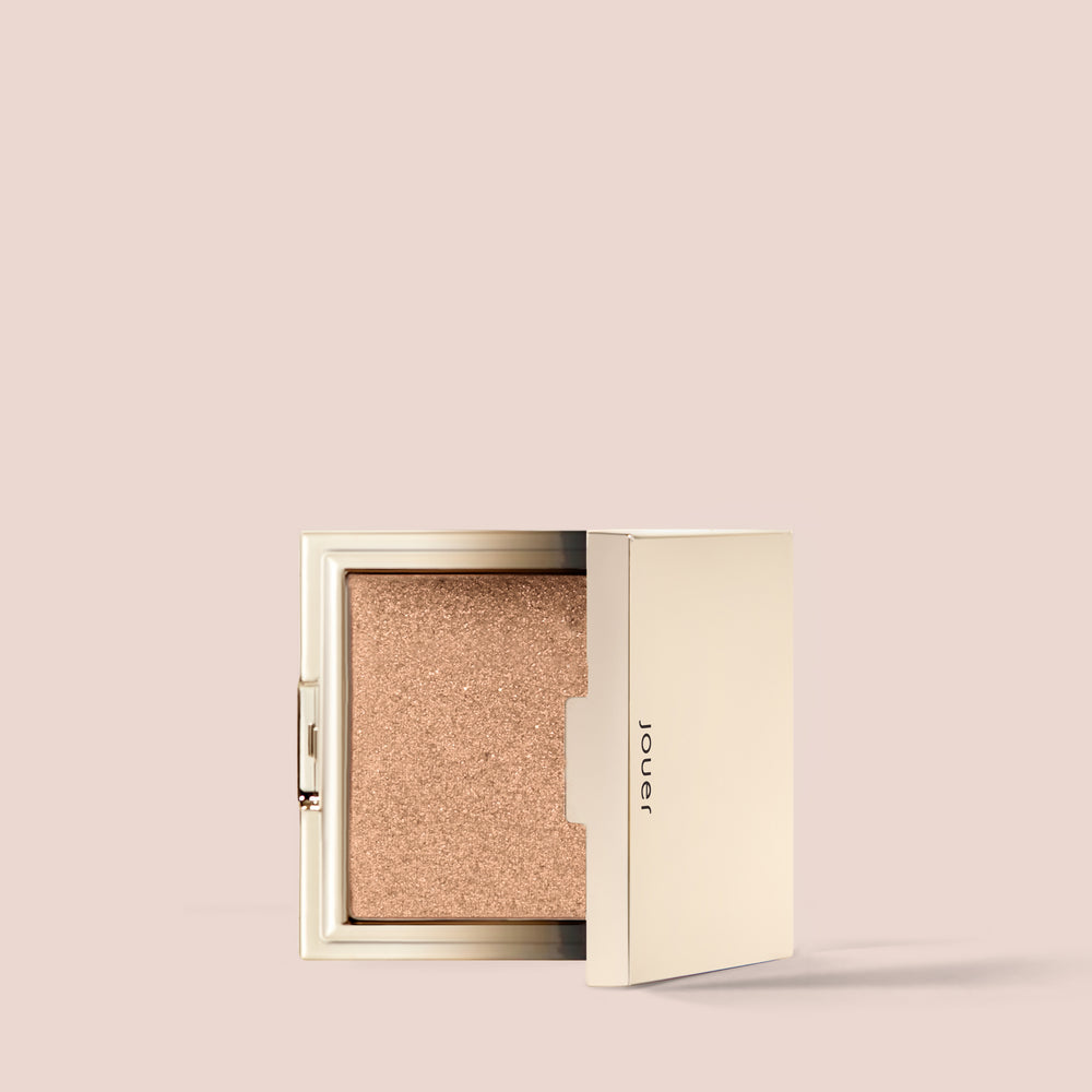 Skinny dip, A sleek, open compact with a shimmery golden powder inside. The Powder Highlighter compact boasts a minimalistic design, featuring clean lines and a soft gold exterior. The brand name "Jouer Cosmetics" is subtly embossed on the side of the cruelty-free, vegan compact. The background is a pale, neutral shade.