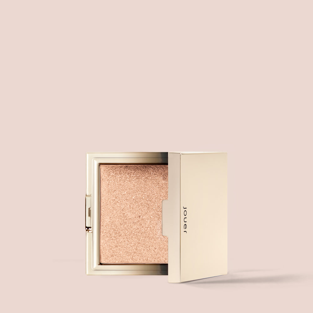 Against a plain, light beige background, an open compact case reveals a shimmering beige highlighter powder inside. The gold-colored compact, designed with minimalist elegance, prominently features the word "Jouer" on the inside of the lid. This Powder Highlighter by Jouer Cosmetics is vegan and cruelty-free, offering radiance without compromise.