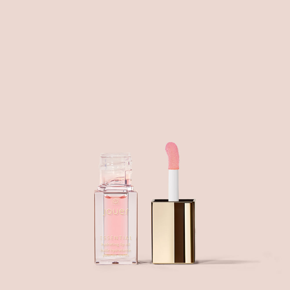 An open tube of Essential Lip Oil from Jouer Cosmetics, featuring a golden lid, is displayed against a plain beige background. The vegan lip oil contains pink-tinted liquid, and the applicator wand has a soft pink sponge tip. The transparent, square-shaped packaging showcases the luxurious product within.