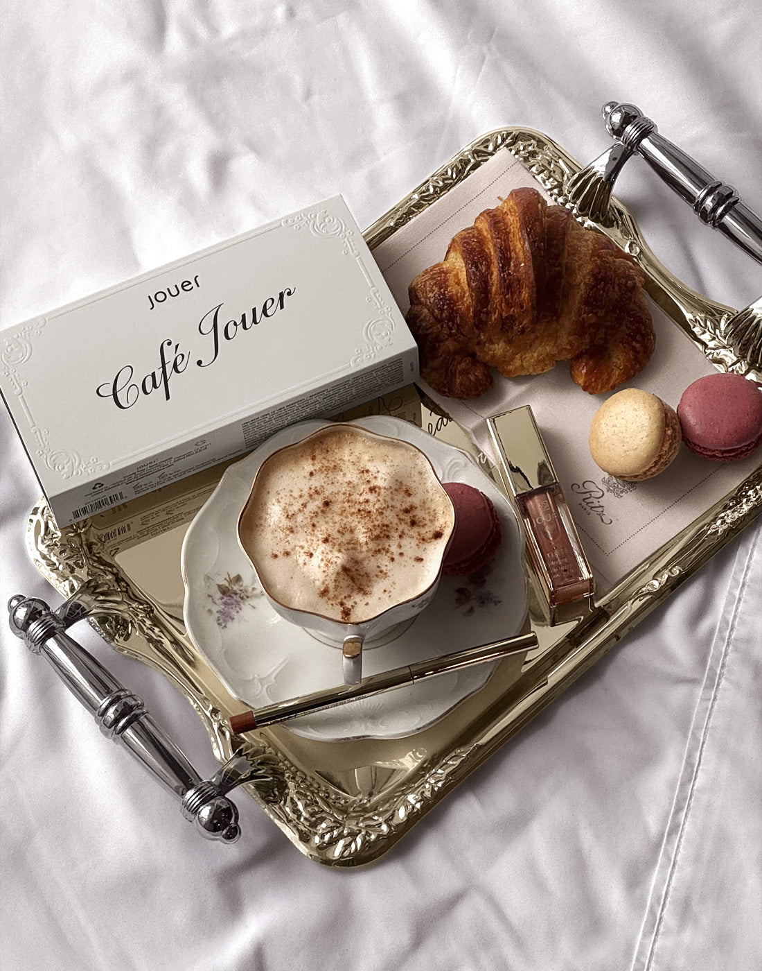 Jouer Café Jouer makeup collection displayed on an elegant gold tray with a cappuccino, croissant, macarons, and lip gloss.