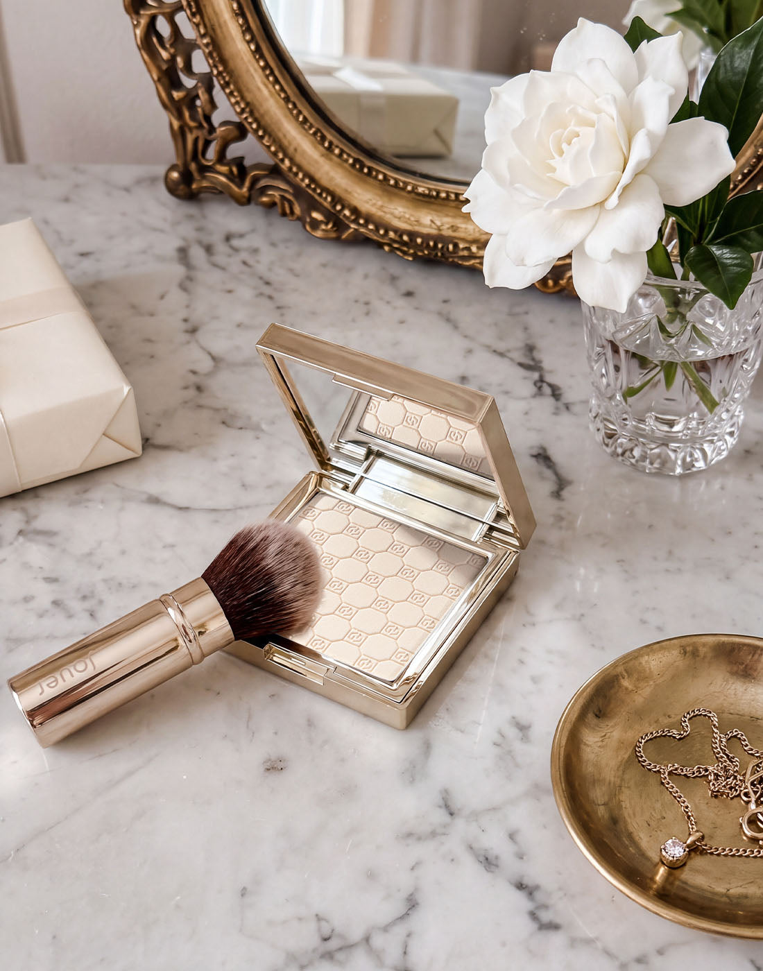 Jouer Soft Focus Powder in a gold compact with a makeup brush, displayed on a marble vanity alongside white gardenias, gold jewelry, and elegant accessories.