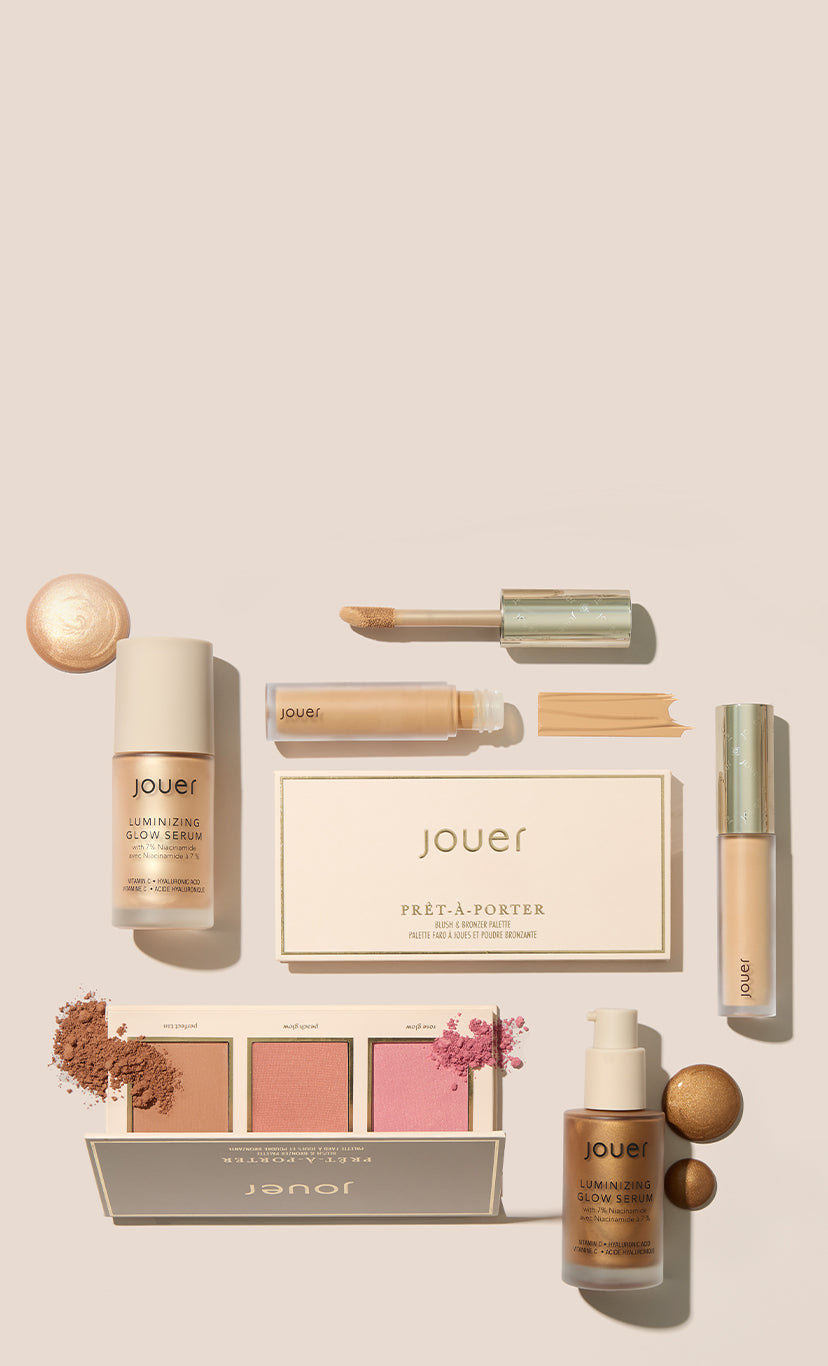 Jouer holiday makeup products on a blush backdrop, including lip gloss, blush, highlighter, and powder compacts with gold packaging.