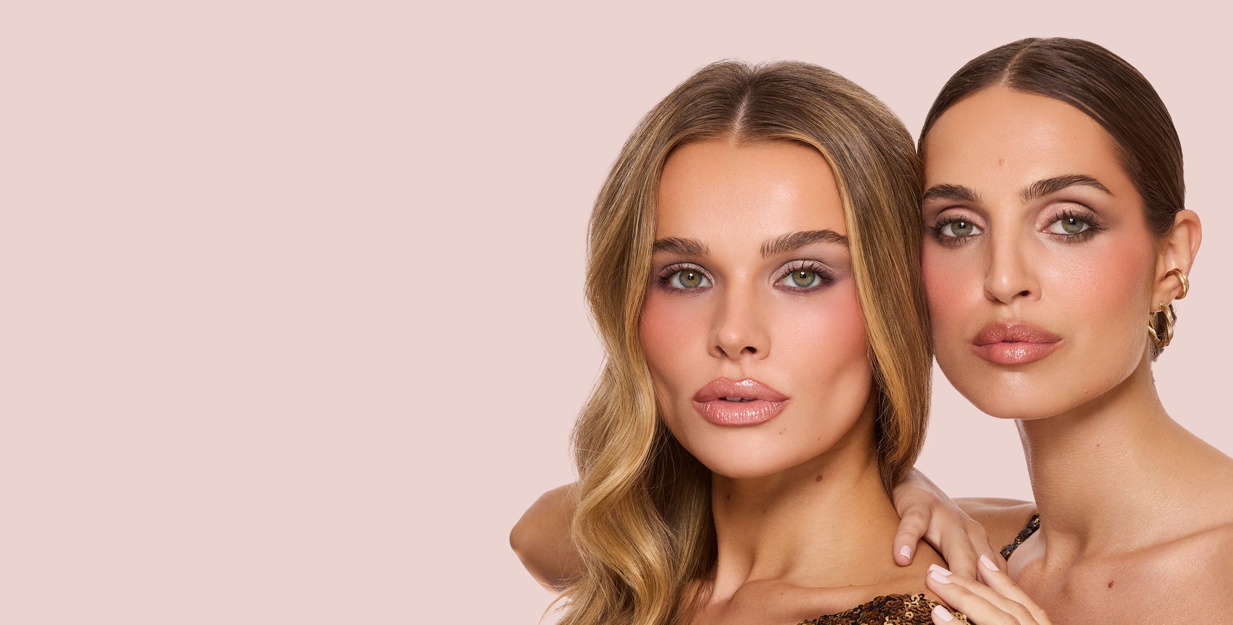 Two models wearing Jouer Cosmetics holiday collection makeup on the right side of a blush background—one in a gold sequin top, the other with slicked-back hair and gold hoop earrings.