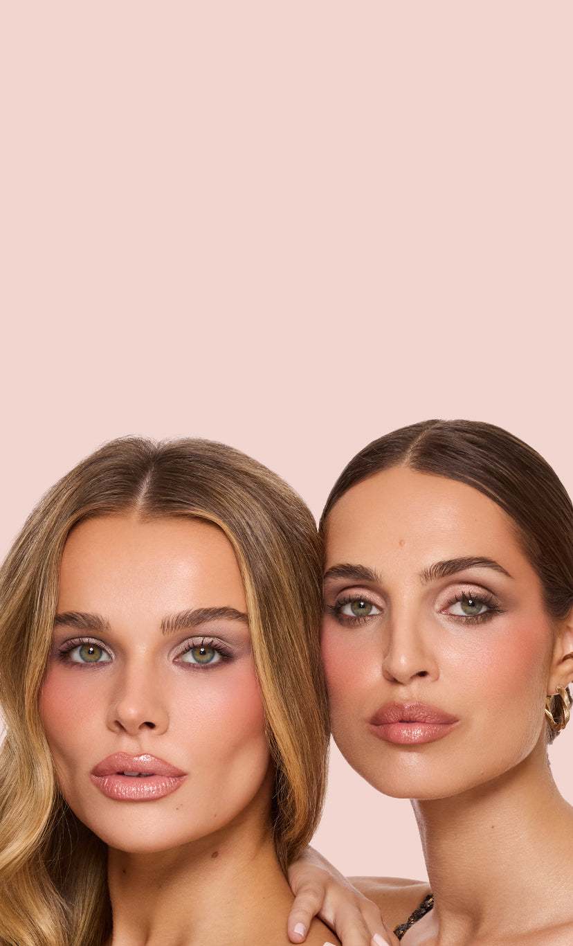 Two models wearing Jouer Cosmetics holiday collection makeup on the right side of a blush background—one in a gold sequin top, the other with slicked-back hair and gold hoop earrings.