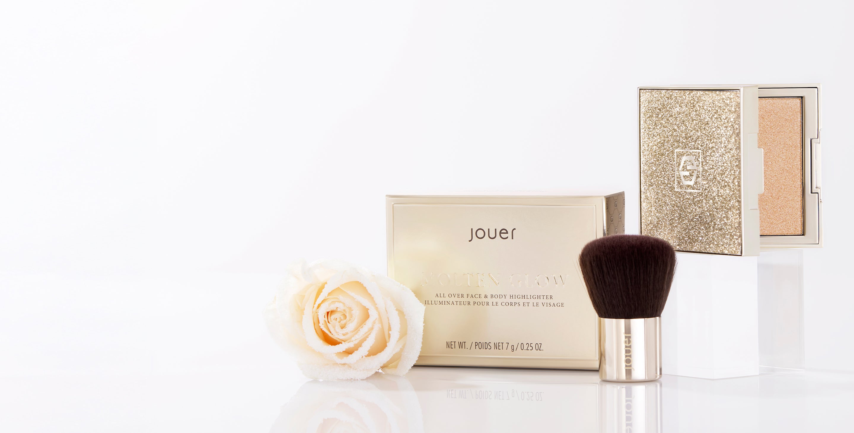 Jouer Molten Glow Highlighter in a glittery gold compact displayed with its box, a soft makeup brush, and a white rose on a clean white background.