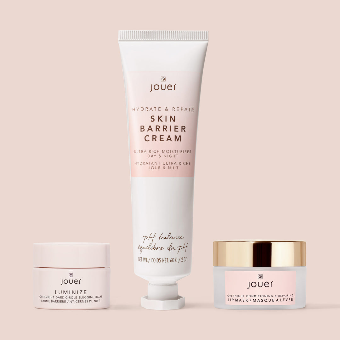 The Bedtime Beauty Set from Jouer Cosmetics, featuring three skincare products, is elegantly arranged against a beige background. This cruelty-free and vegan collection includes a Skin Barrier Cream in a tube, Luminize Overnight Brightening Balm in a jar, and an Overnight Conditioning & Repairing Lip Mask also in a jar.