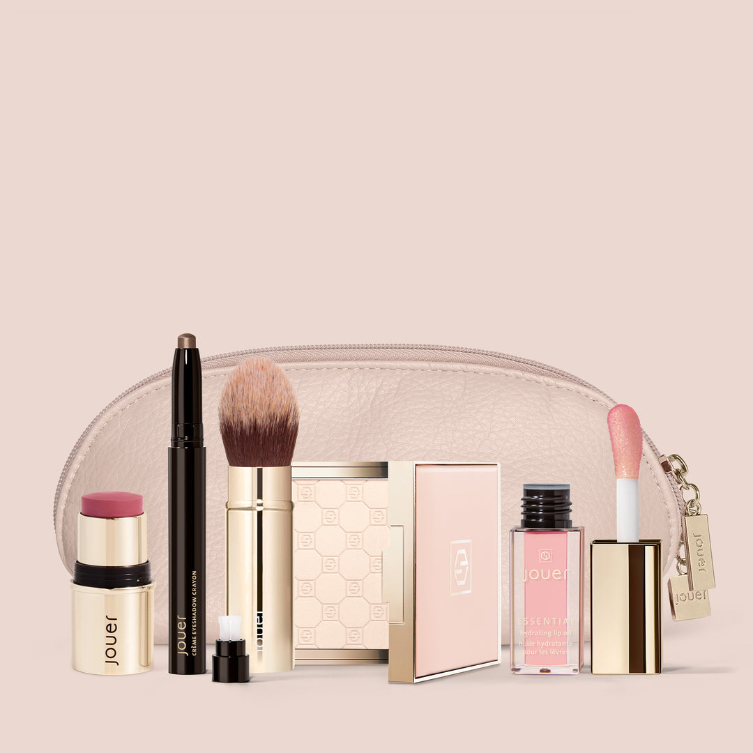 A collection of Jouer Cosmetics' 5 Minute Face products is arranged in front of a beige makeup bag. The items include a blush stick, highlighting stick, makeup brush, pencil eyeliner, compact powder with mirror, lip gloss, and lipstick—all in elegant gold and pink packaging. Each product is Cruelty Free.