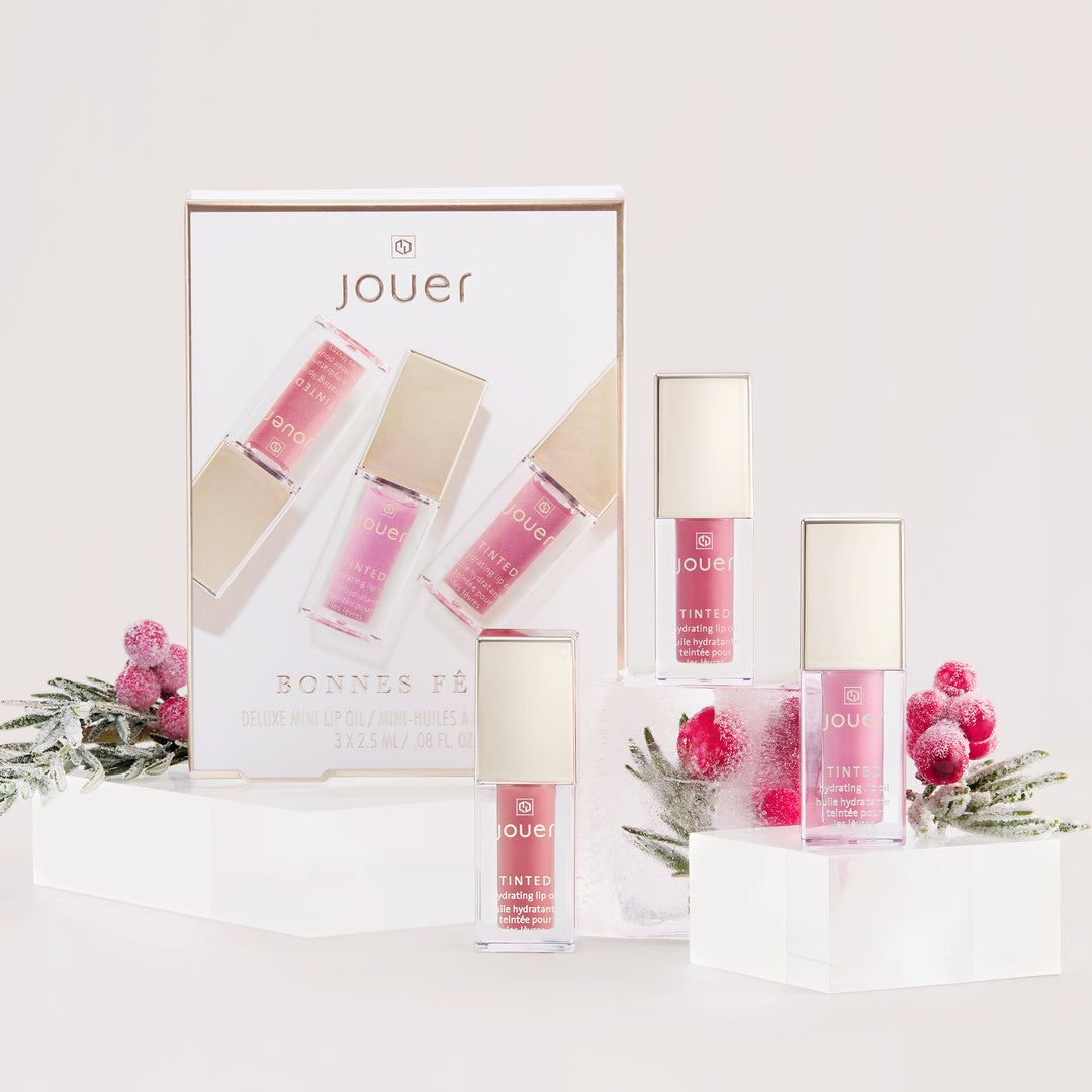 Introducing the Bonnes Fêtes cosmetics set by Jouer Cosmetics, featuring deluxe mini lip glosses. The beautiful packaging displays four vibrant gloss shades, with one gloss standing next to the box, surrounded by festive decorations of frosted greenery and red berries on elegant white rectangular stands. These luxurious glosses are both cruelty-free and paraben-free.