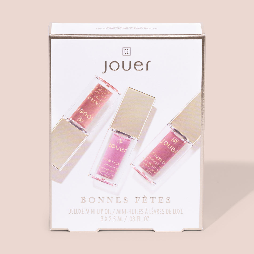 A box of Jouer Cosmetics' "Bonnes Fêtes" Deluxe Mini Lip Oil set contains three mini lip oil bottles in shades of pink and coral. Each bottle, holding 2.5 ml, is topped with a reflective gold cap and displayed in a diagonal arrangement. The lip oils are both paraben-free and cruelty-free.