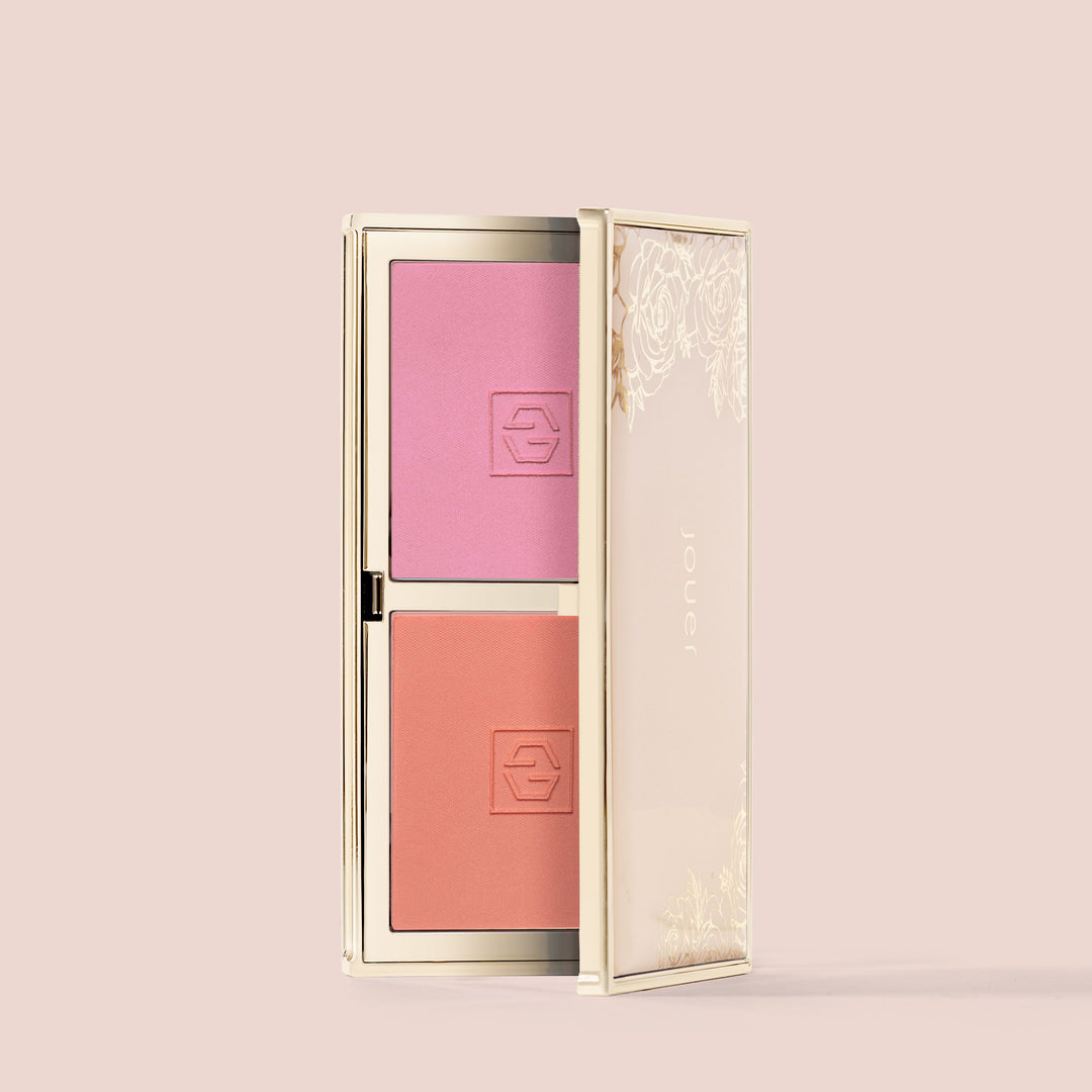 Deux Amis, An open makeup compact, Blush Bouquet by Jouer Cosmetics, features two shades of blush: a pink hue on the top and a coral hue on the bottom. The compact has a gold trim and a decorative floral design on the semi-transparent lid. The word "Jouer" appears on the right side of the lid, highlighting its cruelty-free, vegan formula.