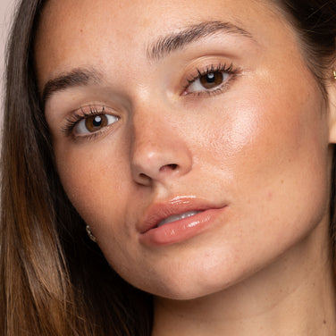  Close-up of a person with long brown hair and smooth, glowing skin. They have brown eyes, natural makeup, glossy lips, and are looking directly at the camera with a neutral expression. Their beauty routine features Jouer Cosmetics' Powder Highlighter to maintain such flawless, radiant skin using cruelty-free products.