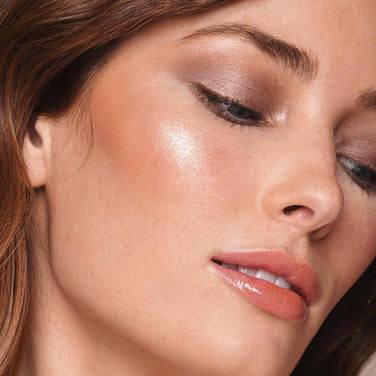  Close-up of a light-skinned individual with brown hair, showcasing a vegan makeup look. They have shimmery eyeshadow, winged eyeliner, glossy pink lips, and glowing highlighter on their cheeks thanks to the Powder Highlighter by Jouer Cosmetics. Their expression is serene as they gaze downwards.