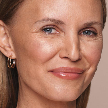 Close-up of a smiling person with light skin, blue eyes, and blonde hair. They are adorned with Jouer Cosmetics' Powder Highlighter for a natural glow and are wearing gold hoop earrings. The background is a soft beige.