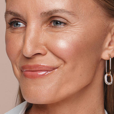  Close-up of a woman smiling. She has light skin, blue eyes, and light brown hair. Her lips are adorned with a vegan, glossy pink lipstick. She wears sparkling hoop earrings shaped like elongated ovals with small crystals. The natural lighting beautifully accents her skin's texture, enhanced by Jouer Cosmetics' Powder Highlighter.