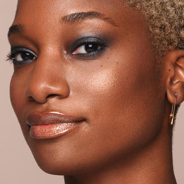  Close-up of a person with short, curly hair who has glowing skin, wearing shimmery eye shadow, glossy lip makeup, and small hoop earrings with a charm. The background is neutral, enhancing the focus on their radiant complexion accentuated by Jouer Cosmetics' Powder Highlighter, which is both vegan and cruelty-free.