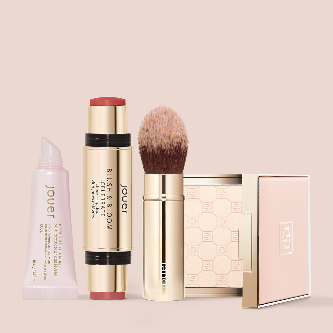 Jouer Essential Lip Enhancer, Blush & Bloom Cheek + Lip Duo, fluffy face brush, and an open Soft Focus powder compact arranged in a row on a soft blush pink background