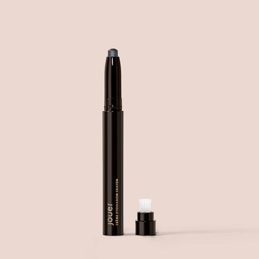  A Crème Eyeshadow pencil in Marble from Jouer Cosmetics, featuring a detachable cap, is set against a beige background. This cruelty-free pencil has a dark tip designed to define your eyelid, and it includes a white applicator positioned near the base next to the cap.