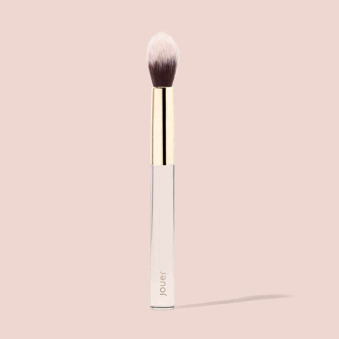 The Jouer Cosmetics Essential Powder Brush is a cruelty-free makeup brush with a gold ferrule and a sleek white handle, featuring soft, rounded bristles that transition from dark to light. The word "Jouer" is printed on the handle. The brush is set against a light beige background.