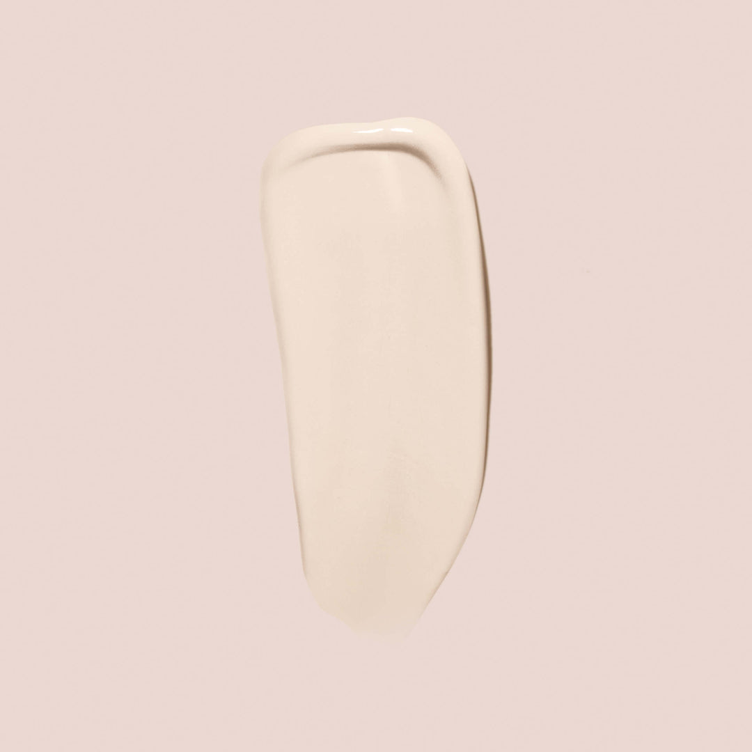 alabaster, full size alt: A close-up of a smear of Essential Crème Foundation by Jouer Cosmetics on a light beige background. The smooth and slightly glossy texture suggests that this paraben-free product is likely a luxurious cosmetic foundation. The elongated shape with rounded edges further highlights its creamy consistency.
