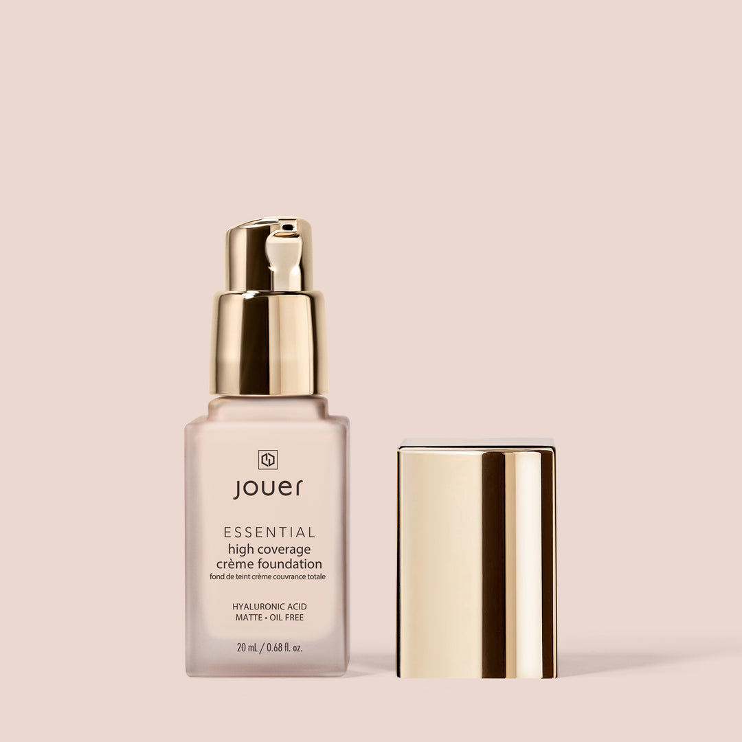 ivory, full size alt:A bottle of Jouer Cosmetics Essential Crème Foundation featuring a pump dispenser and a metallic gold cap placed beside it. The label highlights the inclusion of hyaluronic acid and its matte, oil-free formula, while also proudly stating that it is gluten-free and cruelty-free. The background is a solid light beige color.