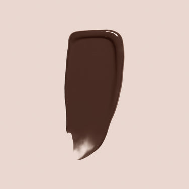  A smear of Jouer Cosmetics' Essential Crème Foundation, showcasing a glossy dark brown hue and free from cruelty and parabens, is spread unevenly on a light beige background. The smear features slightly jagged edges and a tapered end.