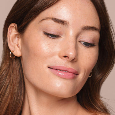  A woman with long brown hair and fair skin is looking down and slightly smiling. She is wearing small, gold hoop earrings and has minimal makeup, giving her a natural look that was likely achieved with Jouer Cosmetics' *Essential Liquid Concealer*. Her smooth skin has a slight glow against the beige background.