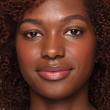  Close-up image of a person with a warm brown complexion, curly hair, and subtle makeup, using an *Essential Liquid Concealer* brush by Jouer Cosmetics to achieve natural eyebrows, light pink blush, and light pink lipstick. The individual has a small nose piercing on their left nostril and a serene, gentle expression.