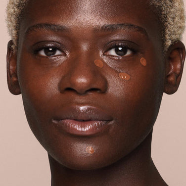  A person with dark skin and short blonde hair applies several dots of *Essential Liquid Concealer from Jouer Cosmetics on their cheeks and chin using a vegan concealer brush. The background is beige.