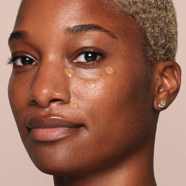  A person with short, blonde hair and a neutral expression looks at the camera with several small dots of Jouer Cosmetics' Essential Liquid Concealer applied under their right eye using a concealer brush. They wear a small, square-shaped earring in their right ear against a plain beige background.