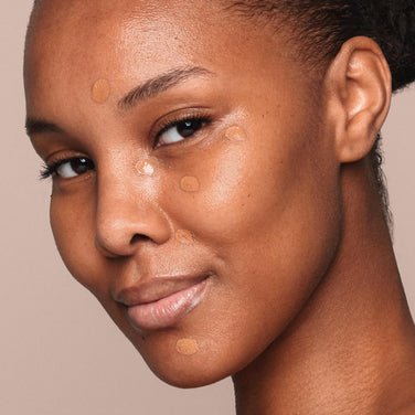  A woman with a smooth complexion and a subtle smile stands against a neutral background. She has several small dots of beige, paraben-free *Essential Liquid Concealer from Jouer Cosmetics applied to her cheeks, forehead, and chin, appearing in the process of blending them in with a vegan concealer brush.