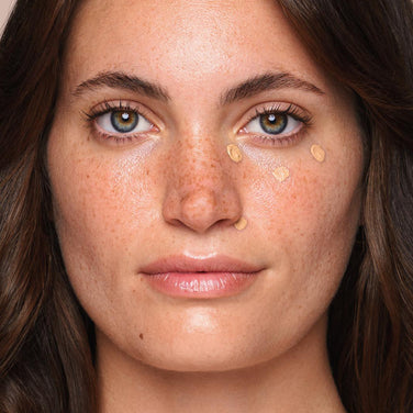  A close-up of a young woman with fair skin and freckles applying the *Essential Liquid Concealer* from Jouer Cosmetics under her eyes. She has green eyes, long brown hair, and slightly parted lips. The creamy, paraben-free concealer is dotted on her cheeks just below her eyes as she uses a concealer brush. The background is light and blurred.
