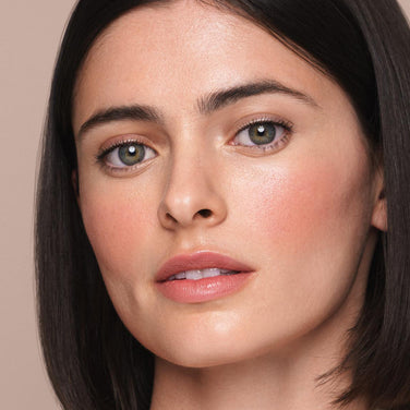  Close-up of a person with medium-length dark hair and a light complexion, looking slightly to the side. They have well-groomed eyebrows, blush on their cheeks, and natural-looking makeup applied with the *Essential Liquid Concealer from Jouer Cosmetics. The background is beige.