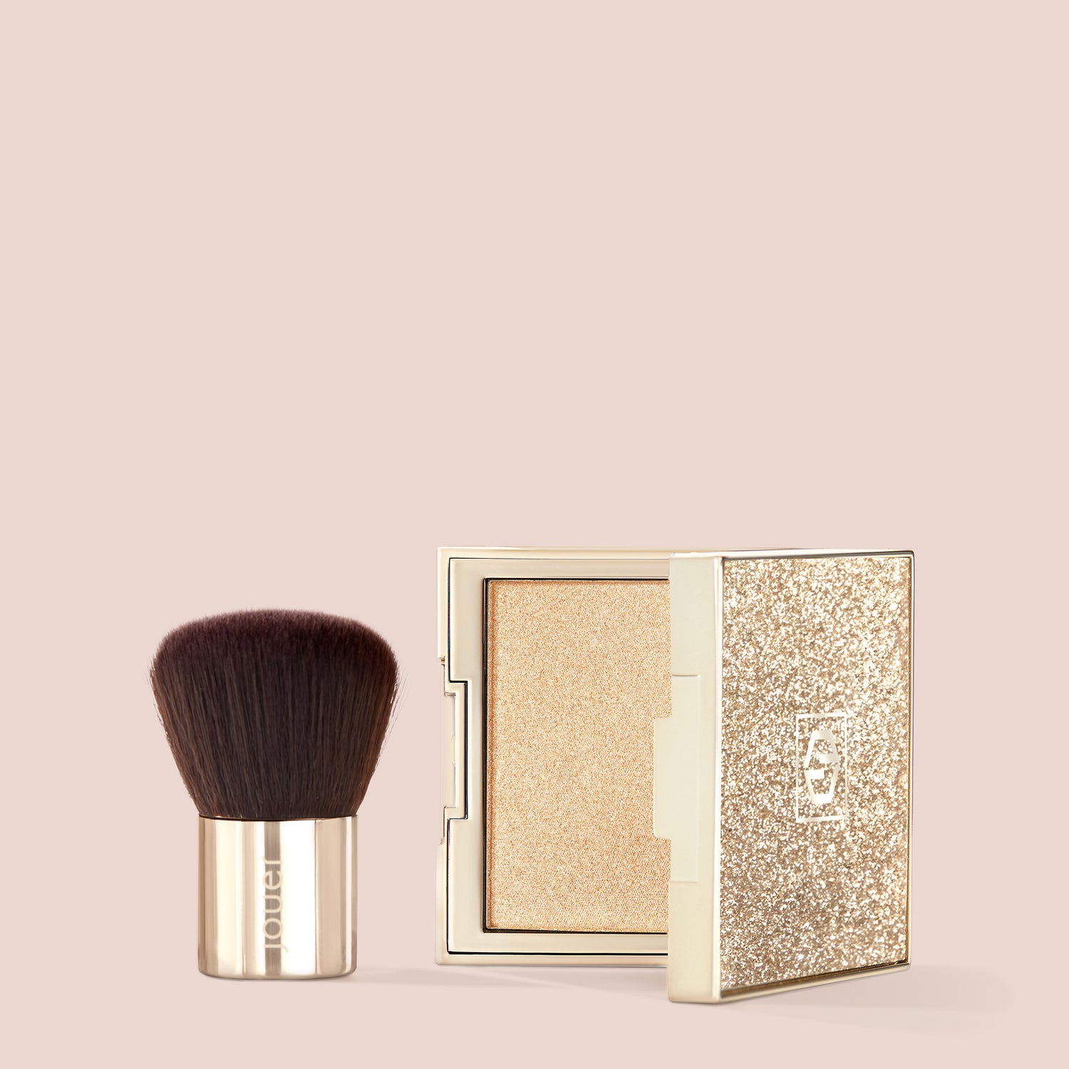 A compact named "Molten Glow" by Jouer Cosmetics, featuring a sparkling gold lid and slightly open to unveil a luxurious highlighter, sits beside a Petit Kabuki Brush with dense brown bristles. Together, they promise to deliver a luminous glow against the beige background.