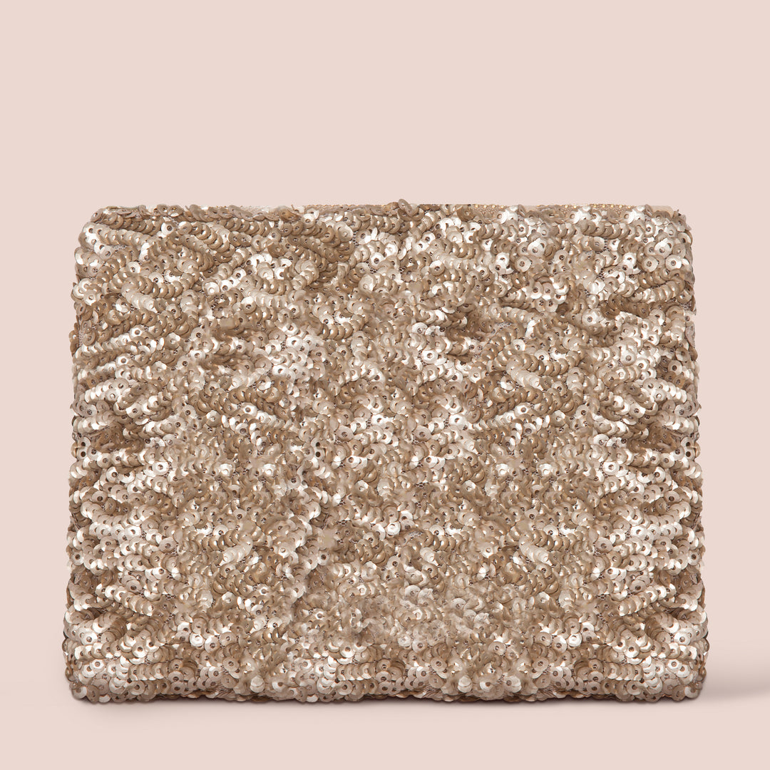 The Gold Sequins Bag by Jouer Cosmetics is a rectangular clutch embellished with shimmering gold sequins, offering a textured and sparkling surface. It features a discreet zipper closure and is set against a plain, light beige background, adding an elegant touch to any ensemble.